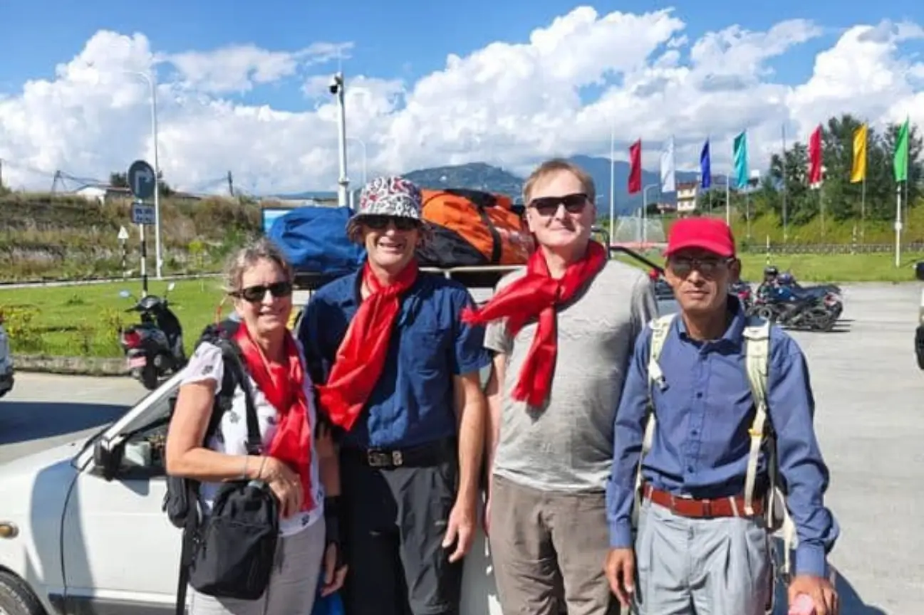 Four smiling trekkers in matching red scarves posing with their guide beside a car in a mountain town, ready for their journey.