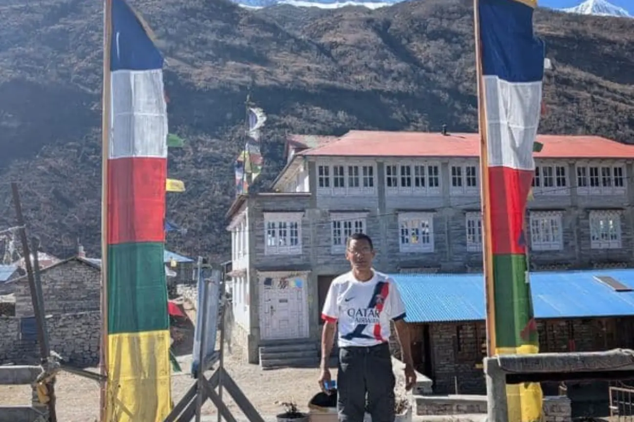 A trekker standing under a traditional wooden village archway adorned with colorful prayer flags, with a stone house and mountains behind him.