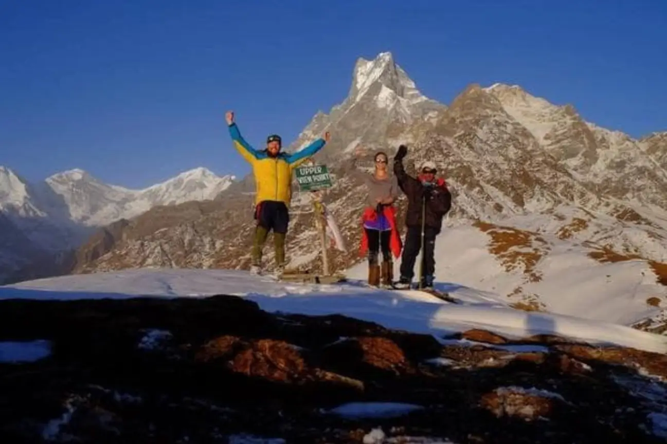 Three ecstatic trekkers at a snowy high-altitude viewpoint with arms raised, celebrating reaching a summit with the iconic Machhapuchhre peak in the background.