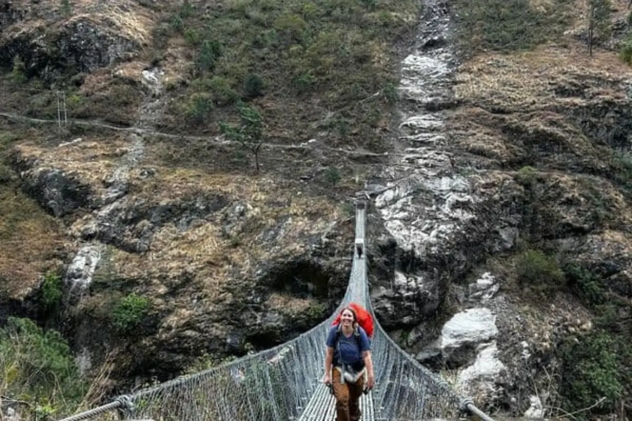 A smiling female trekker with a red backpack carefully crossing a long, high suspension bridge over a deep, rocky river gorge.