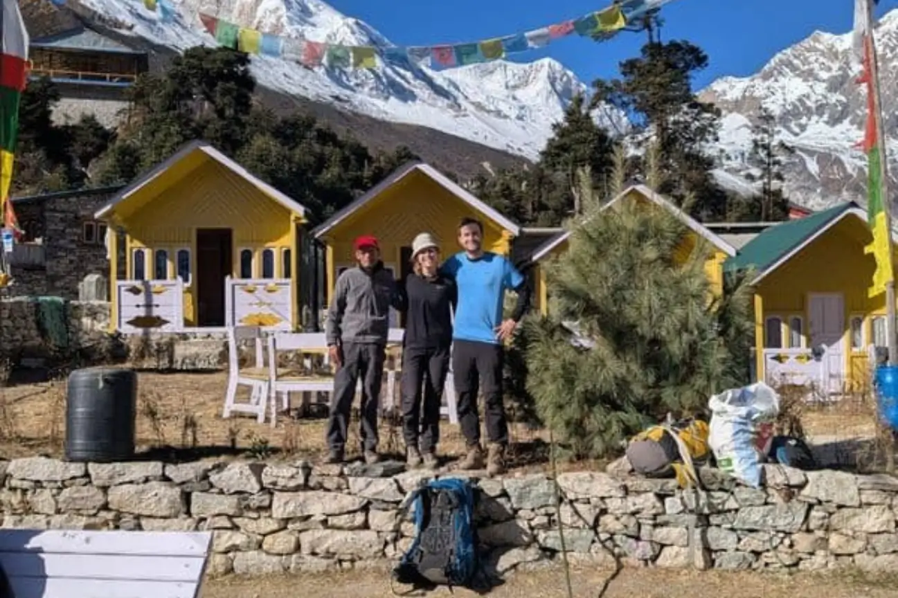 A trekking group posing in front of cozy, yellow teahouse lodges with stunning snow-capped mountain views and prayer flags.